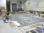 工作室一角的工作情形。工作人員正作在地上將石塊一片片拼貼至底稿上；由此可見要完成一幅巨型馬賽克的工程何其浩大。